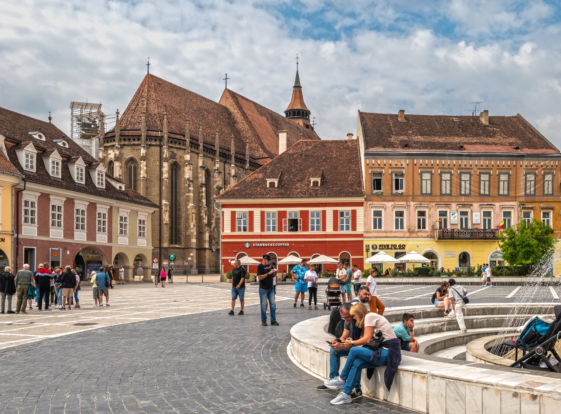 Junge Leute entspannen am Brunnen auf dem Rathausplatz in Brașov, dahinter die Schwarze Kirche